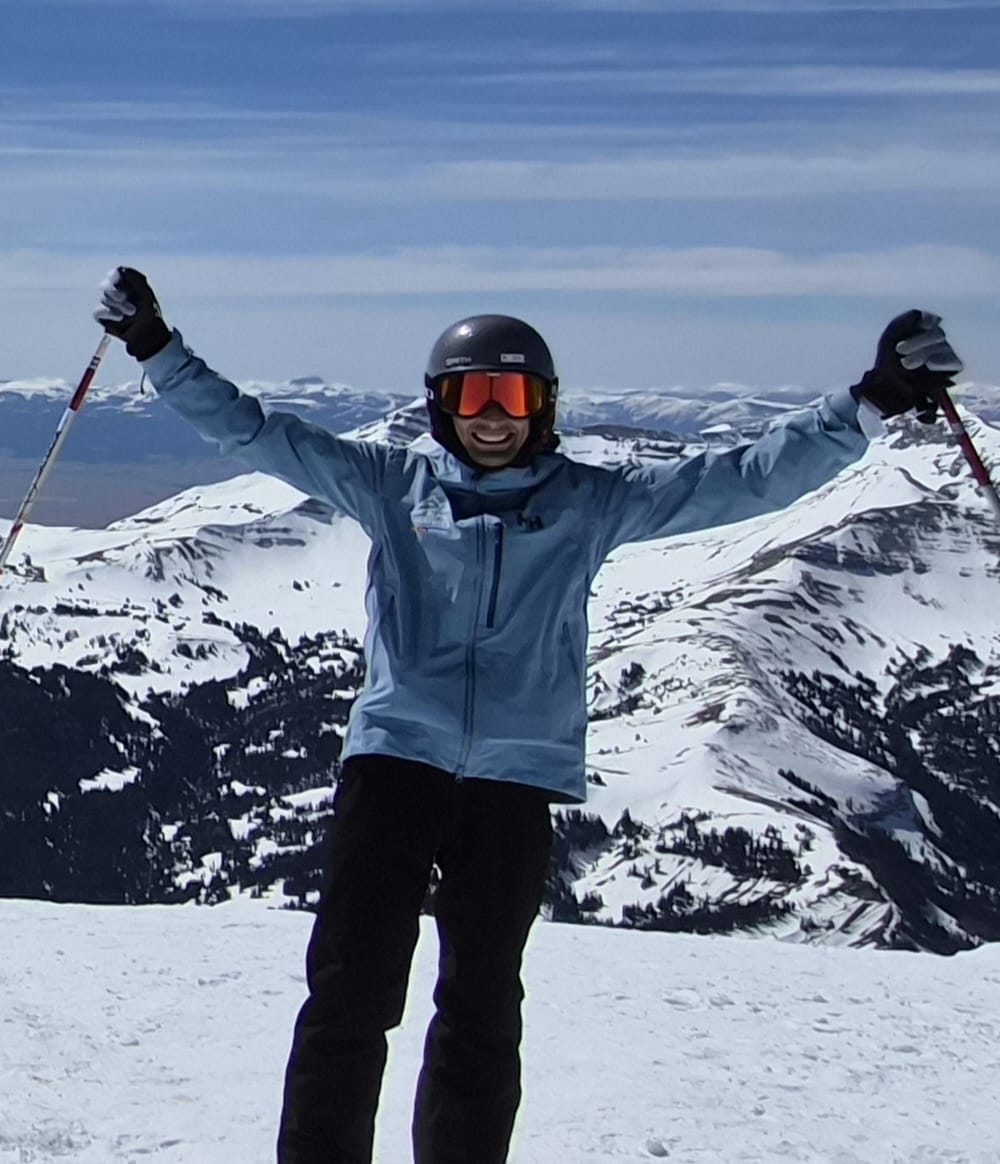 Octávio de braços abertos, com bastões de ski, no topo de uma montanha nevada sob céu azul